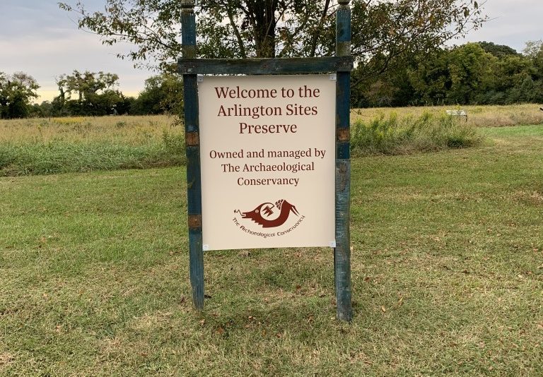 Photo of an information placard installed in a green field. The placard reads "Welcome to Arlington Sites Preserve. Owned and Managed by the Archaeological Conservancy"