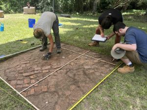 Three people are bending over to examine an excavated square of earth where there are bricks that have been exposed. One person is picking something up and another is drawing on a notepad.