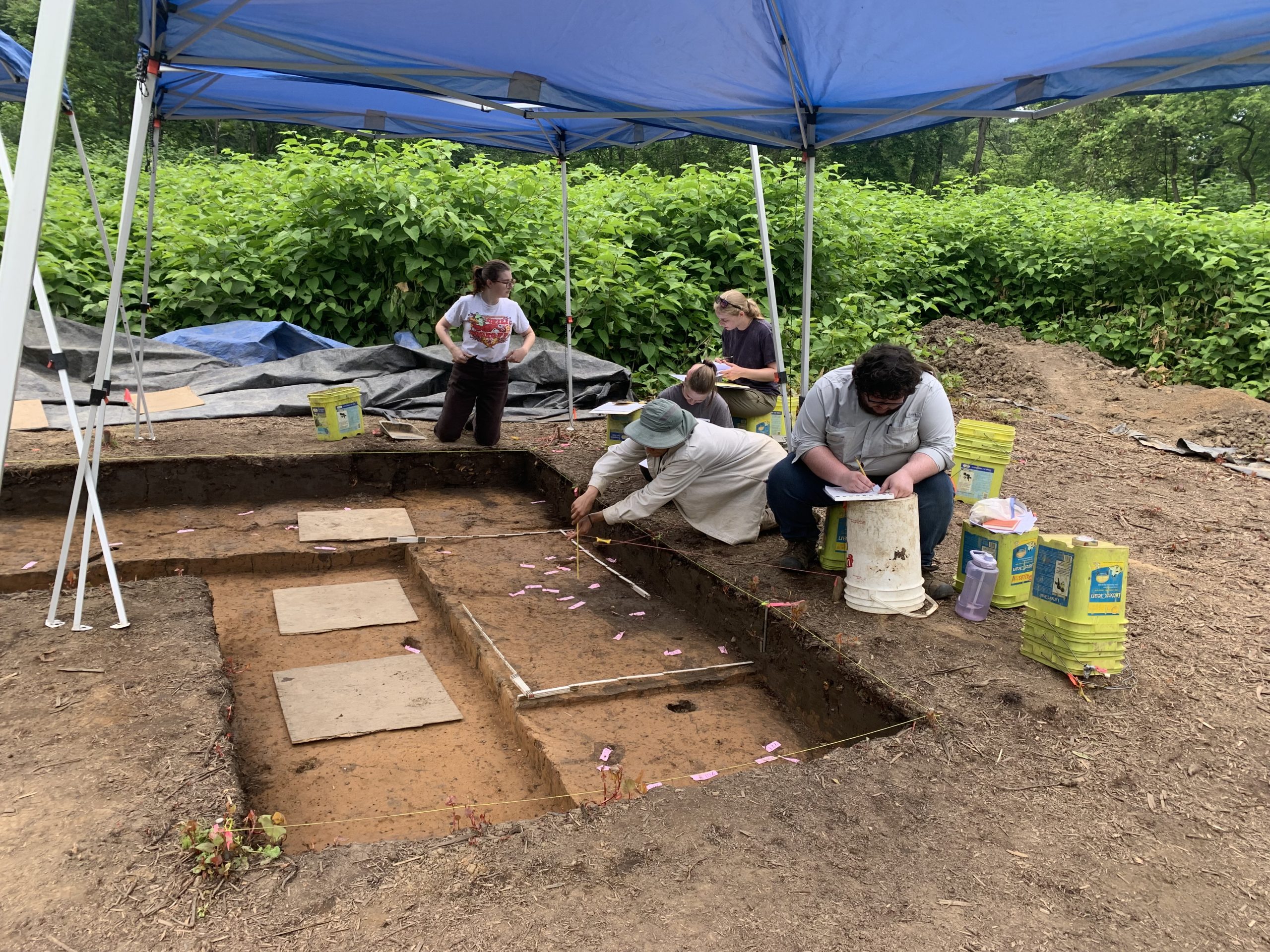 Photo of four people sitting under a blue awning looking over an open archaeological excavation. Several are taking notes.